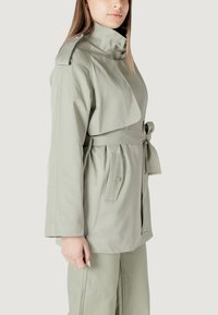 Cappotto trench verde chiaro con colletto a risvolto, cintura in vita e tasche laterali. Presenta una texture liscia e maniche lunghe con polsini.