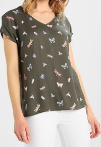 Olivgrünes V-Ausschnitt-Shirt mit Druck von pinken, blauen und weißen Schmetterlingen, Libellen und Käfern, getragen mit weißen Hosen.