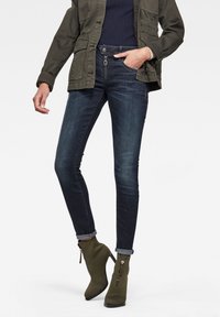 Jeans ajustados en denim oscuro con una textura suave, que presentan un cierre con cremallera en el frente y puños doblados, combinados con botas hasta el tobillo en color verde oliva.