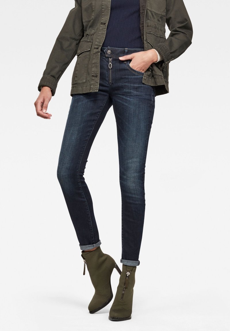 Jeans ajustados en denim oscuro con una textura suave, que presentan un cierre con cremallera en el frente y puños doblados, combinados con botas hasta el tobillo en color verde oliva.