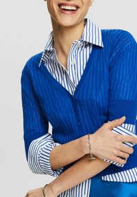 Modrý žebrovaný cardigan přes bílou košili s modrými vertikálními pruhy. Má výstřih do V, zapínání na knoflíky a na zápěstí stříbrný náramek.