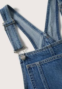 Gros plan d'une salopette en denim bleu avec bretelle ajustable et boucle métallique sur fond blanc.