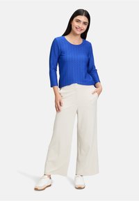 Blau strukturierter Langarmshirt mit rundem Ausschnitt, kombiniert mit weißen weiten Hosen und weißen Sneakers, vor einem neutralen Hintergrund getragen.