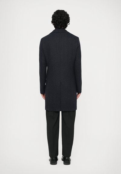 Hombre con cabello rizado que lleva un abrigo largo oscuro, pantalones negros y zapatos negros, de pie de espaldas contra un fondo simple.
