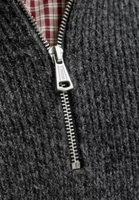 Gros plan d'une fermeture éclair argentée sur un tissu tricoté gris foncé partiellement ouverte pour révéler une chemise à carreaux rouge et blanche en dessous.