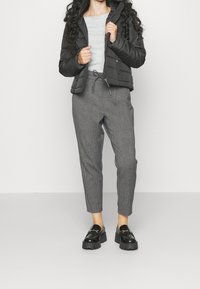 Svart vadderad pufferjacka, grå långärmad tröja, grå byxor i rutmönster med tapered passform, och kraftiga svarta loafers med guldfästen.