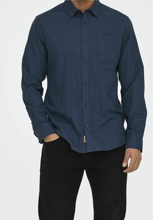 Uomo che indossa una camicia di colore blu scuro con maniche lunghe e taschino sul petto, pantaloni neri, mano in tasca, in piedi su sfondo bianco.