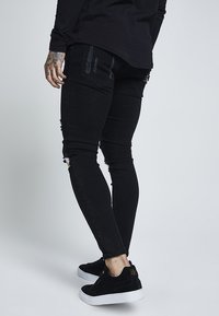 Black skinny jeans with distressed detail, featuring ripped areas at the knees and textured, faded patches. Paired with black sneakers.