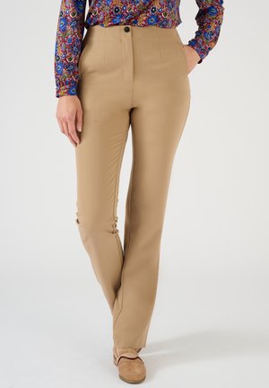 Femme portant un pantalon beige avec les mains dans les poches, associé à une chemise à manches longues à fleurs colorées et des mocassins beiges.