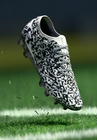 Čierno-biele futbalové kopačky s pixelovým vzorom, majú textúrovaný zvršok a výrazné kolíky, zobrazené nad trávou.