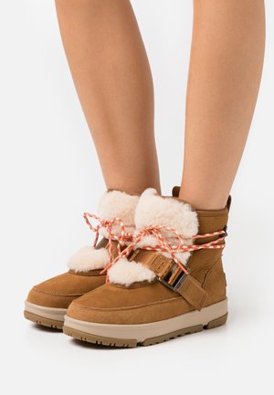 Bottes d'hiver en daim marron avec doublure en polaire blanche, lacets orange et blancs, boucle latérale, portées sur des jambes nues sur fond blanc.