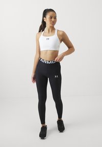 Hvit sport BH med brede stropper og logo, kombinert med høy midje svarte leggings som har "ARMOUR" på midjebandet og svarte sneakers.