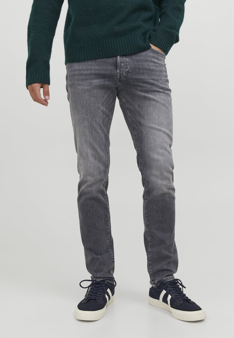 Jeans de mezclilla gris con ajuste slim, que presentan un acabado desgastado. Combinados con zapatillas azul marino y un suéter de punto verde.