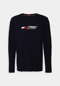 Pitkähihainen tummansininen puuvillapaita, jossa on valkoinen ja punainen vaakasuuntainen logo rinnassa. Pyöreä kaula-aukko, jossa raidallinen sisus.