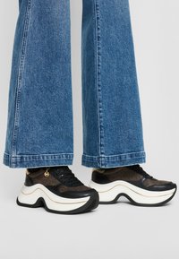 Ténis pretos e castanhos com sola branca espessa, apresentando um design texturizado, detalhes metálicos e sola exterior distinta em forma de onda.