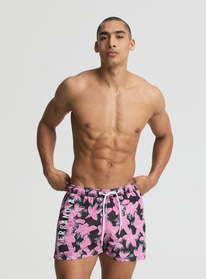 Uomo muscoloso che sistema dei pantaloncini da bagno neri con grande stampa floreale rosa e coulisse bianca su uno sfondo chiaro e uniforme.