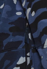 Zip-up hoodie med ett blått camouflage-mönster i varierande nyanser, texturerat tyg och en svart dragkedja med en tygflik.