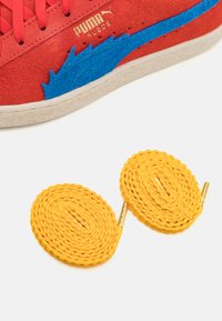 Røde ruskinds sneakers med blå filt detaljer, præsenterer en tekstureret overflade og guld logo. Inkluderer to gule runde snørebånd med metalspidser.