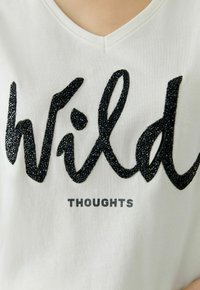 Weißes V-Ausschnitt-T-Shirt mit schwarzem strukturiertem Schriftzug "Wild" und funkelnden Akzenten sowie grauem "THOUGHTS" darunter. Weiches Baumwollmaterial.