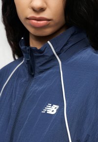 Το ελαφρύ μπουφάν σε ναυτικό μπλε χρώμα διαθέτει ψηλό γιακά, λευκή διακοσμητική γραμμή και μικρό λευκό λογότυπο New Balance στο στήθος. Υφή υφάσματος.