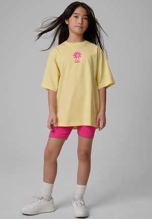 Dziewczyna o długich ciemnych włosach, ubrana w luźny żółty T-shirt z różową grafiką słońca, różowe spodenki, białe skarpetki i białe trampki, stoi na szarym tle.