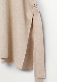Pull en maille beige avec un long détail de lien à la fente latérale, suspendu sur un fond blanc.