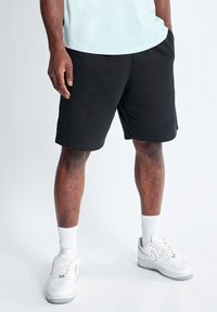 Pantalones cortos de algodón negros con bolsillos laterales, cinturilla elástica y corte relajado, combinados con zapatillas blancas y calcetines de canalé hasta el tobillo.