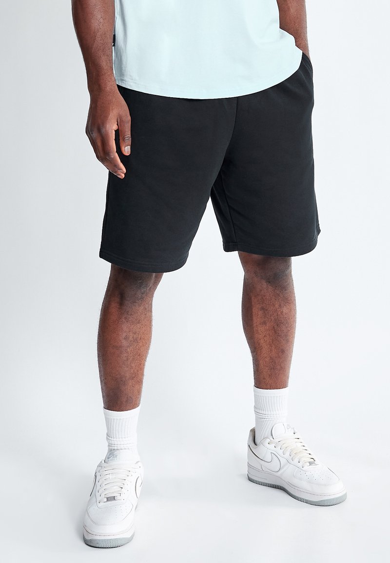 Pantalones cortos de algodón negros con bolsillos laterales, cinturilla elástica y corte relajado, combinados con zapatillas blancas y calcetines de canalé hasta el tobillo.