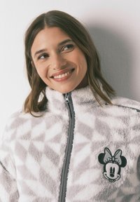 White fleece jacket with a geometric pattern in light gray, featuring a black Minnie Mouse patch on the left chest and a front zipper.