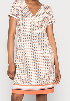 Kobieta ubrana w sukienkę z krótkim rękawem i dekoltem w serek, z geometrycznym wzorem w kolorach pomarańczowym, czarnym i białym, z paskowym obszyciem w tych samych kolorach.