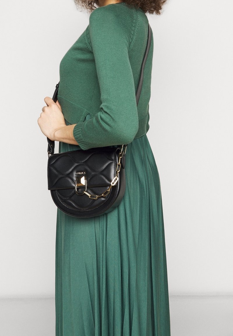 Donna in abito verde a maniche lunghe che tiene una borsa a sella nera trapuntata con catena e chiusura in oro sulla spalla, davanti a uno sfondo semplice.