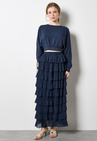 Marineblaues zweiteiliges Outfit mit einem bauchfreien Langarmoberteil und einem gestuften, bodenlangen Rock mit plissierten Lagen. Sandalen mit dünnen Riemen.