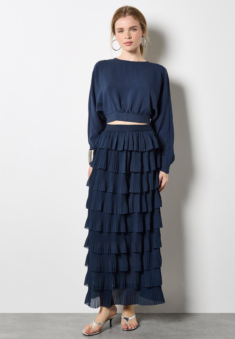 Marineblaues zweiteiliges Outfit mit einem bauchfreien Langarmoberteil und einem gestuften, bodenlangen Rock mit plissierten Lagen. Sandalen mit dünnen Riemen.