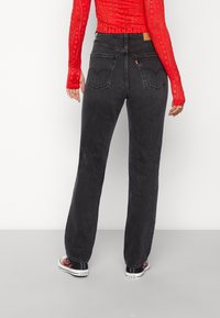 Jeans de denim negro con corte recto, con bolsillos traseros y una etiqueta de marca de cuero. Combinados con zapatos negros con diseño de llamas.