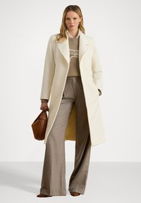 Balts garais mētelis ar izgrieztiem uzgaļiem, bēšs adīts džemperis un pelēkas platas bikses. Aksesuāri iekļauj brūnu rokassomiņu. Viensfonts fons.