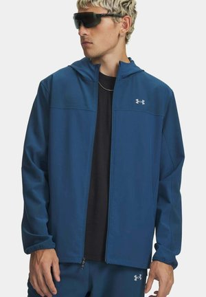 Man met een donkerblauw Under Armour jack met rits en bijpassende broek, zwart T-shirt, zwarte zonnebril en zilveren ketting.