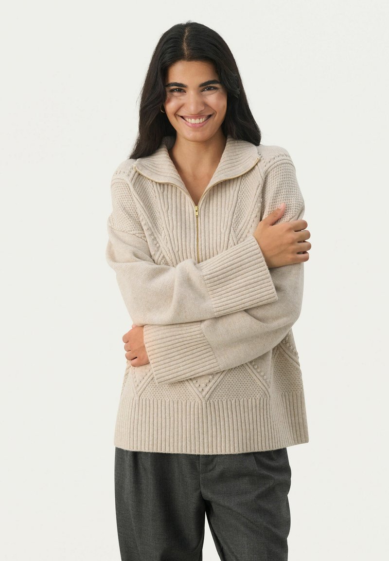 Beige knit sweater with a zip collar, featuring ribbed cuffs and textured patterns. Soft material and oversized fit, paired with dark trousers.