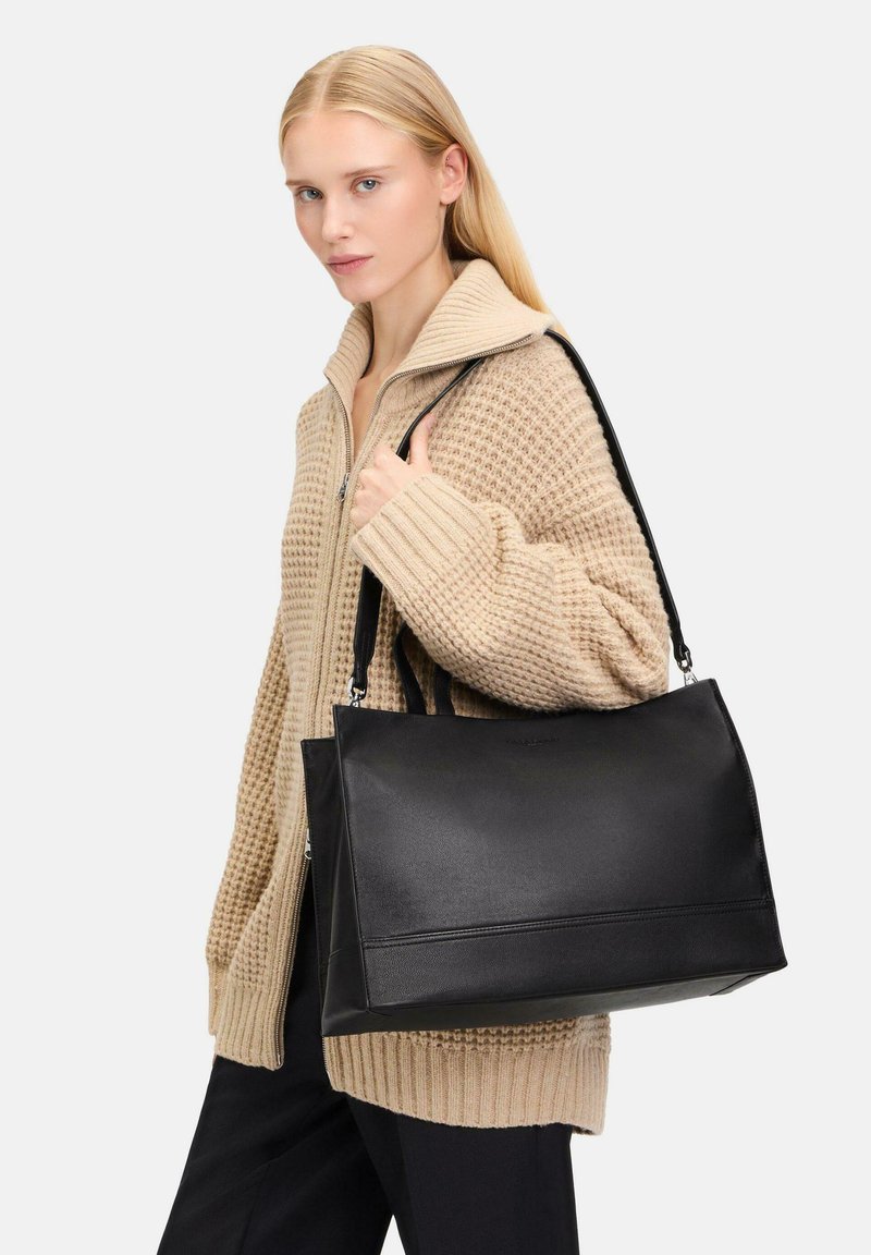 Schwarze Lederhandtasche mit strukturiertem Design und Seitennähten, getragen von einem Model in einer beigen Strickjacke.