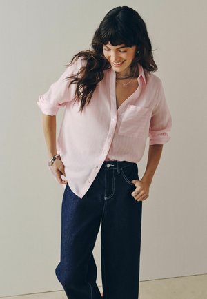 Frau lächelt und blickt nach unten, trägt ein hellrosa Hemd mit Knöpfen und hochgekrempelten Ärmeln sowie dunkle Jeans mit weitem Bein, steht vor einem schlichten Hintergrund.