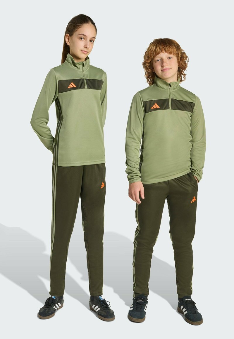 Olivově zelené teplákové soupravy s černými akcenty, zipovým límcem a oranžovým logem; odpovídající kalhoty mají boční pruhy a žebrovanou strukturu.