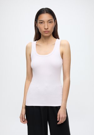 Femme aux cheveux raides et foncés portant un débardeur blanc sans manches et un pantalon noir, debout devant un fond clair uni.