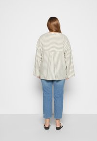 Camisa oversized bege e branca com riscas, com mangas largas, combinada com jeans azul claro e sandálias pretas. Tecido texturizado, corte descontraído.