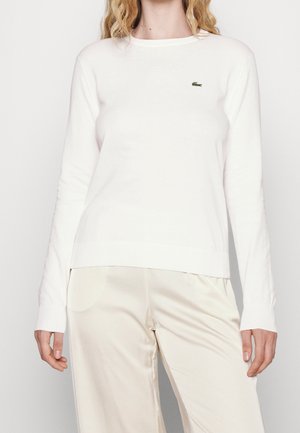 Kobieta ubrana w prostą białą koszulę z długim rękawem z małym zielonym logo krokodyla na lewej piersi oraz jasnobeżowe spodnie.