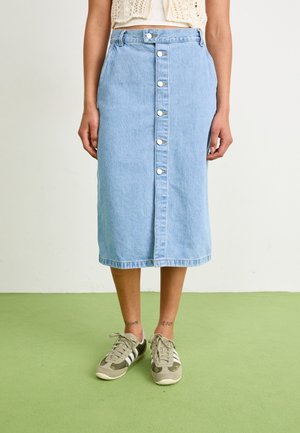 Jupe midi en denim bleu clair avec boutons sur le devant, portée avec des baskets beige et blanches sur un sol vert, raccourcie au-dessus de la taille.