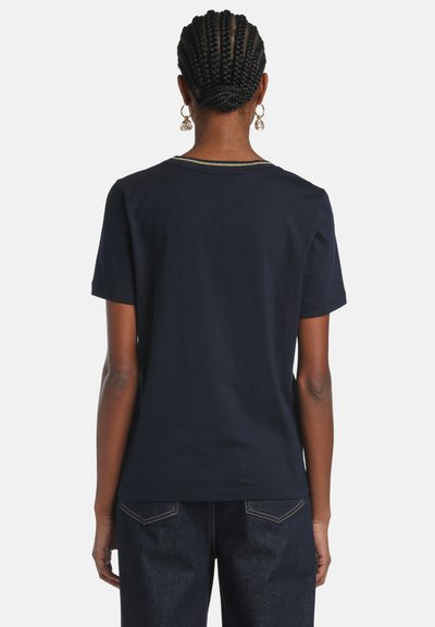 Fonott hajú és karika fülbevalót viselő személy, egyszínű sötét tengerészkék rövid ujjú ingben és sötét farmernadrágban, háttal a nézőnek.