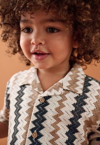 Kurzärmliges Hemd mit Knopfleiste in beigen, braunen und schwarzen Zickzackmustern. Mit Kragen und holzähnlichen Knöpfen. Strukturierter Stoff.