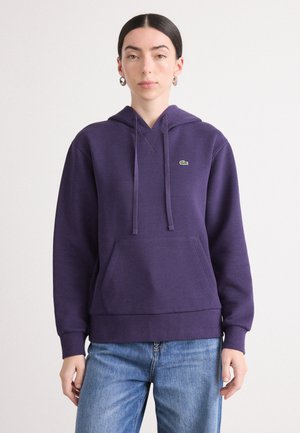 Lilla hettegenser laget av myk stoff, med en frontlomme og en liten grønn logo på brystet. Løs passform og ribbestrikkede mansjetter.