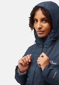 Veste marine imperméable avec capuche ajustable, fermeture éclair à l'avant, poignets élastiques et accents de logo. Légère, texture lisse, adaptée à une utilisation en extérieur.
