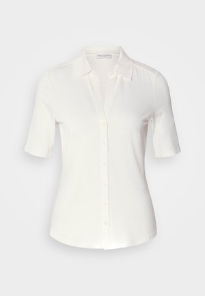 Baltas trumpas rankovėmis mygtukais užsegamas marškinys, pagamintas iš švelnios, lygios medžiagos. Turi klasikinį apykaklę ir aptemptą siluetą su švariu apdaila.