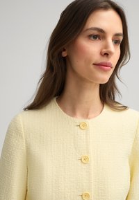 Giacca gialla con texture, caratterizzata da un collo rotondo e tre bottoni gialli sul davanti, che mette in risalto un design in tessuto intrecciato.